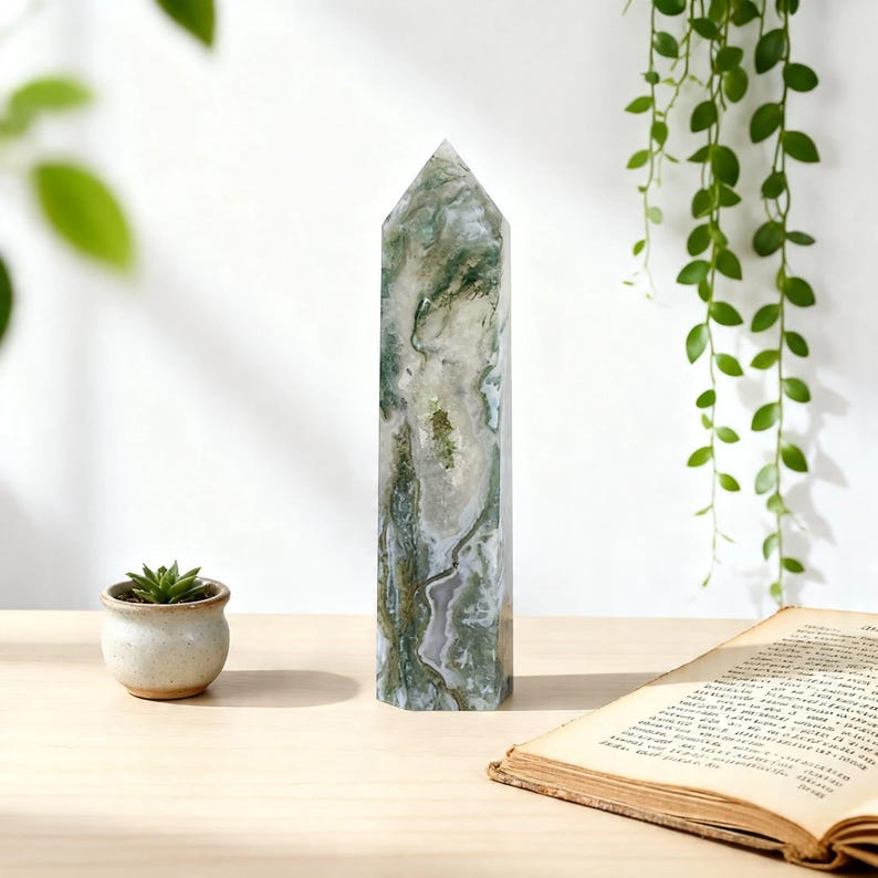 May include: A tall, rectangular crystal obelisk with a pointed top. The crystal features a mix of white, green, and gray colors. A small potted succulent and an open book are also visible on a light-colored wooden surface.