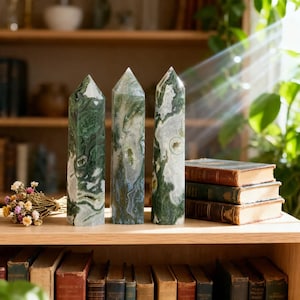 May include: Three tall, polished stone obelisks with green and white patterns stand on a wooden shelf. The obelisks are next to a small bouquet of dried flowers and a stack of vintage books. The background includes a bookshelf and natural light.