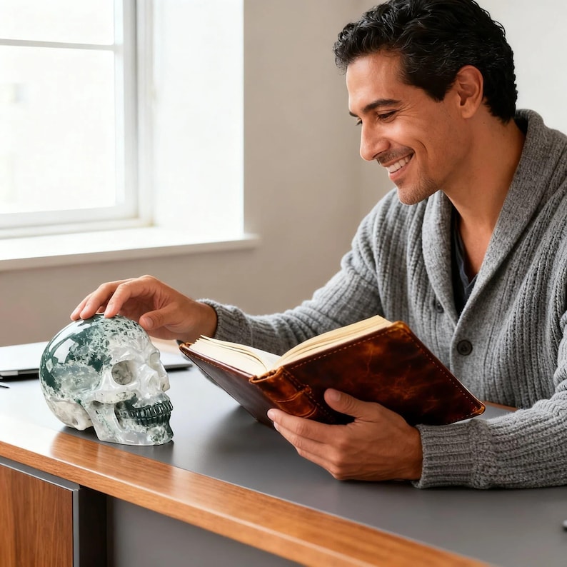 May include: A decorative skull made of green and white stone sits on a desk. The skull is next to a brown leather-bound journal with cream-colored pages. The person in the image is wearing a gray cardigan.