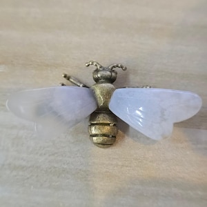 May include: A bee-shaped brooch featuring a textured, antique-style brass body and head. The insect has two large, translucent, pale lavender-colored wings made of smooth, polished stone that extend horizontally from the central metal body.