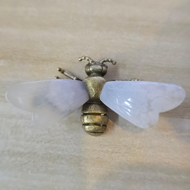 May include: A bee-shaped brooch featuring a textured, antique-style brass body and head. The insect has two large, translucent, pale lavender-colored wings made of smooth, polished stone that extend horizontally from the central metal body.