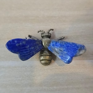 May include: A bee-shaped brooch featuring a metallic bronze-toned body with segmented details and two large, flat wings crafted from deep blue lapis lazuli stone with natural white and grey inclusions, complete with two small antennae on the head.