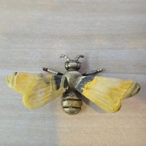 May include: A bee-shaped brooch featuring a textured, antique-style metallic body and two translucent, wing-shaped stones. The wings display a marbled pattern of yellow, cream, and dark gray tones, while the central body is a dull, muted silver.