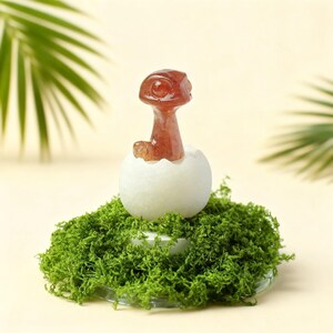 May include: A reddish-brown dinosaur figure emerging from a white egg, set on a bed of green moss. The egg rests on a clear, round base. The background is a soft, neutral colour with green palm leaves.