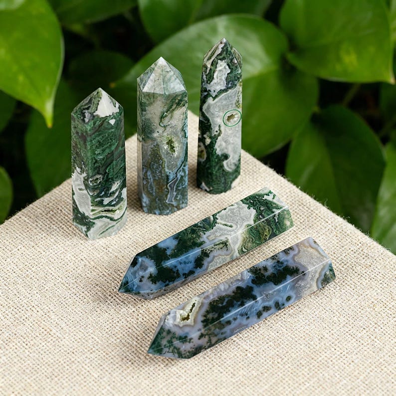 May include: Four green and white moss agate crystal points and obelisks. The crystals have a natural, marbled appearance with shades of green, white, and blue. The crystals are displayed on a beige linen surface.