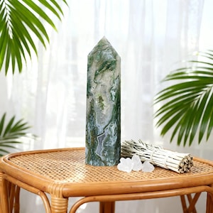 May include: A tall, polished moss agate crystal obelisk with green and white patterns. The crystal is displayed on a woven table, next to a bundle of sage and small clear crystals. Palm leaves are in the background.