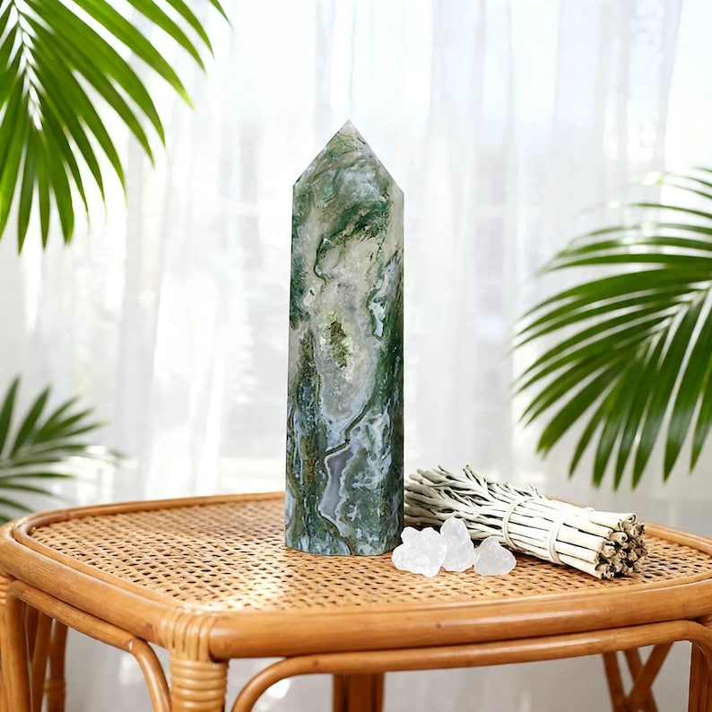 May include: A tall, polished moss agate crystal obelisk with green and white patterns. The crystal is displayed on a woven table, next to a bundle of sage and small clear crystals. Palm leaves are in the background.