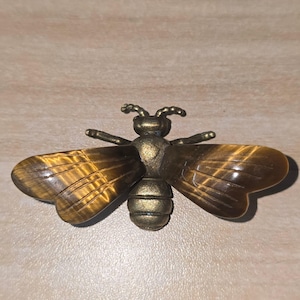 May include: A bee-shaped brooch featuring a metallic bronze-toned body and head with two wings crafted from polished brown and gold tiger's eye gemstone, displaying natural chatoyant banding and carved linear details on the wing surfaces.