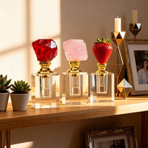 May include: Three decorative perfume bottles with crystal stoppers in red, pink, and a strawberry. The bottles have gold accents and sit on a wooden shelf. A candle holder and framed photo are in the background.