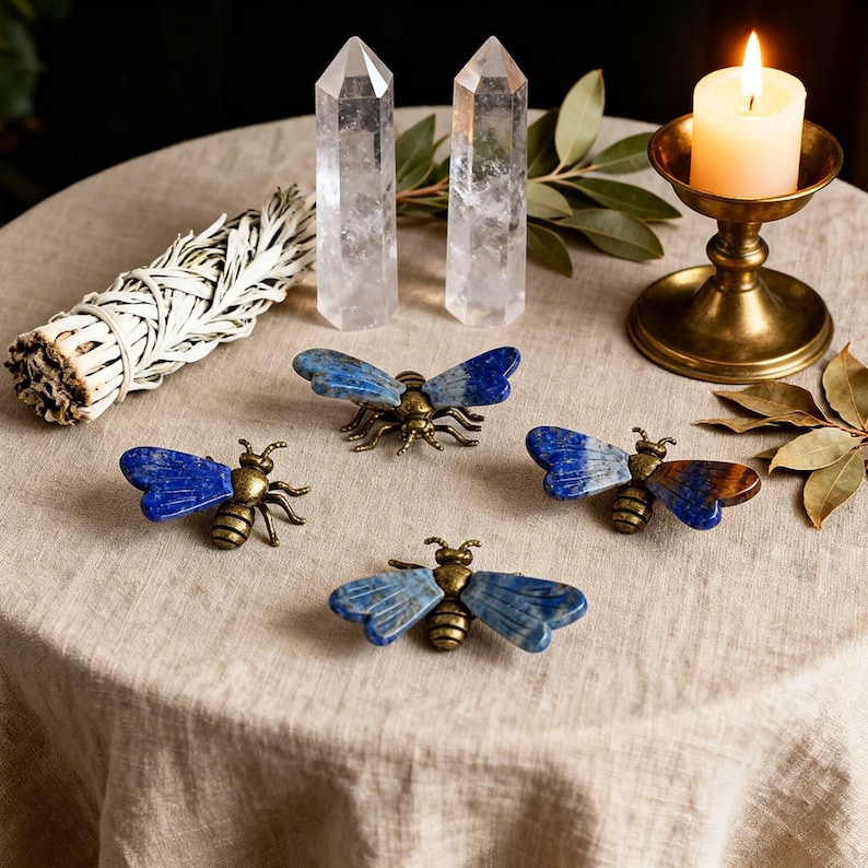 May include: Four bee-shaped ornaments featuring metallic bronze-toned bodies and wings crafted from polished blue lapis lazuli stone. The bees are arranged on a textured surface alongside two clear quartz crystal points and a lit candle.
