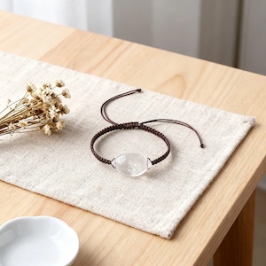 Könnte beinhalten: Ein braunes Makramee-Armband mit einem klaren, oval geformten Kristall. Das Armband liegt auf einem beigen Leinentuch. Im Hintergrund befinden sich getrocknete Blumen und eine kleine weiße Schale. Das Armband ist verstellbar.