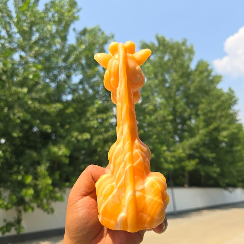 May include: A carved orange and white stone figurine shaped like a giraffe, viewed from the back. The sculpture features a long neck, rounded ears, and a smooth, polished surface with swirling color patterns throughout the mineral structure.
