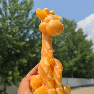 May include: A carved orange stone figurine in the shape of a giraffe. The sculpture features a smooth, polished surface with natural banded patterns in varying shades of amber and gold, depicting the giraffe's head, long neck, and body.