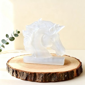 Puede incluir: Una escultura de cabeza de unicornio blanca translúcida con melena y cuerno fluidos. La escultura está montada sobre una base rectangular y se exhibe sobre una rodaja de madera redonda y natural con bordes de corteza.