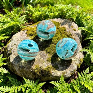 May include: Three spherical blue aragonite crystals with natural cavities and brown mineral inclusions. Each polished stone displays a vibrant turquoise hue with intricate banding and crystalline textures visible within the open geode-like centers.