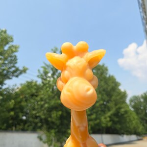 May include: A solid orange giraffe figurine featuring a rounded snout, two small ears, and two ossicones on top of its head. The giraffe has a smooth, molded surface and a long neck, captured in a clear, focused view against an outdoor backdrop.