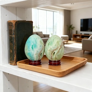 May include: Two egg-shaped decorative stones featuring marbled patterns in shades of teal, mint green, and white. Each stone is mounted on a circular, dark reddish-brown wooden base and sits on a square wooden tray next to a vintage book.