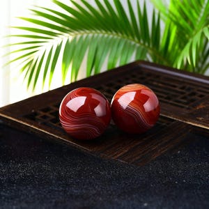 May include: Two polished, red agate spheres with white and brown banding. The spheres are displayed on a dark wooden tray, with green palm fronds in the background. The spheres are approximately 5 cm in diameter.