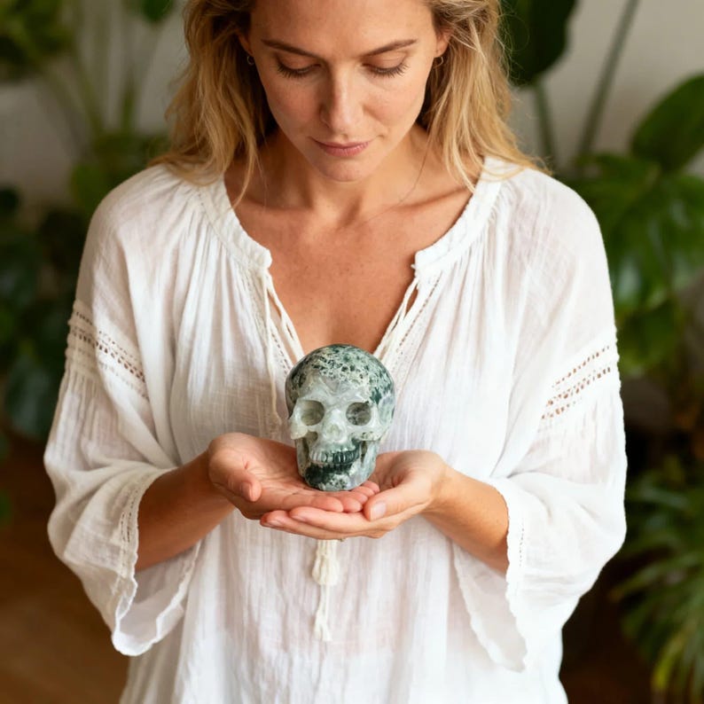 May include: A carved skull made of green and white stone. The skull is held in open hands. The skull has detailed features, including eye sockets, a nose cavity, and teeth. The background is blurred, with green plants visible.