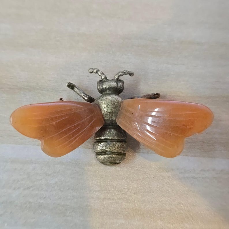 May include: A bee-shaped brooch featuring a textured, antique-style brass-toned metal body and head, paired with two translucent, smooth, orange-colored stone wings that display subtle linear veining across their surface.