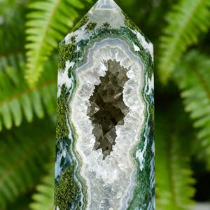 May include: A polished, elongated crystal geode with a hollow center. The crystal features a mossy green and white exterior with a geode cavity lined with sparkling quartz crystals. The background is blurred green foliage.