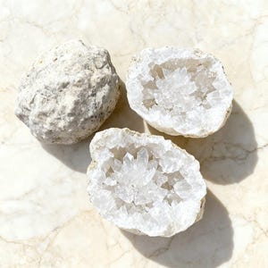 Puede incluir: Tres cristales de geoda blancos. Dos están abiertos, revelando un grupo de cristales de cuarzo transparente en su interior. Las geodas están sobre una superficie de mármol. El exterior de las geodas es áspero y texturizado.