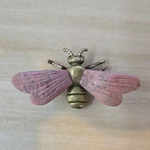 May include: A bee-shaped brooch featuring a metallic bronze-toned body with segmented details and two large, flat wings crafted from smooth, mottled pink stone. The insect design includes small antennae and thin, extended metallic legs.