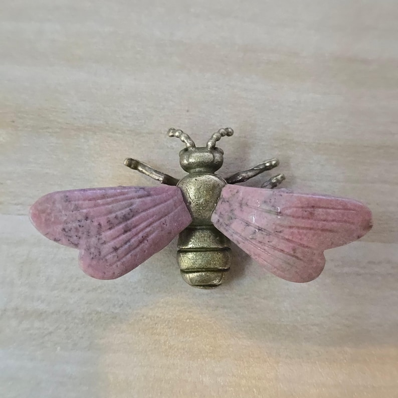 May include: A bee-shaped brooch featuring a metallic bronze-toned body with segmented details and two large, flat wings crafted from smooth, mottled pink stone. The insect design includes small antennae and thin, extended metallic legs.