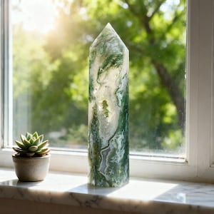 May include: A tall, polished stone obelisk with a pointed top. The stone features a mix of green and white patterns, resembling moss agate. A small potted succulent sits on the windowsill next to the obelisk.