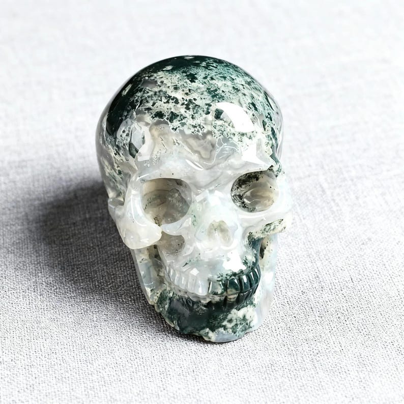 May include: A carved skull made of translucent stone with green and white inclusions. The skull has detailed features, including eye sockets, a nose cavity, and teeth. The stone has a polished, glossy finish.