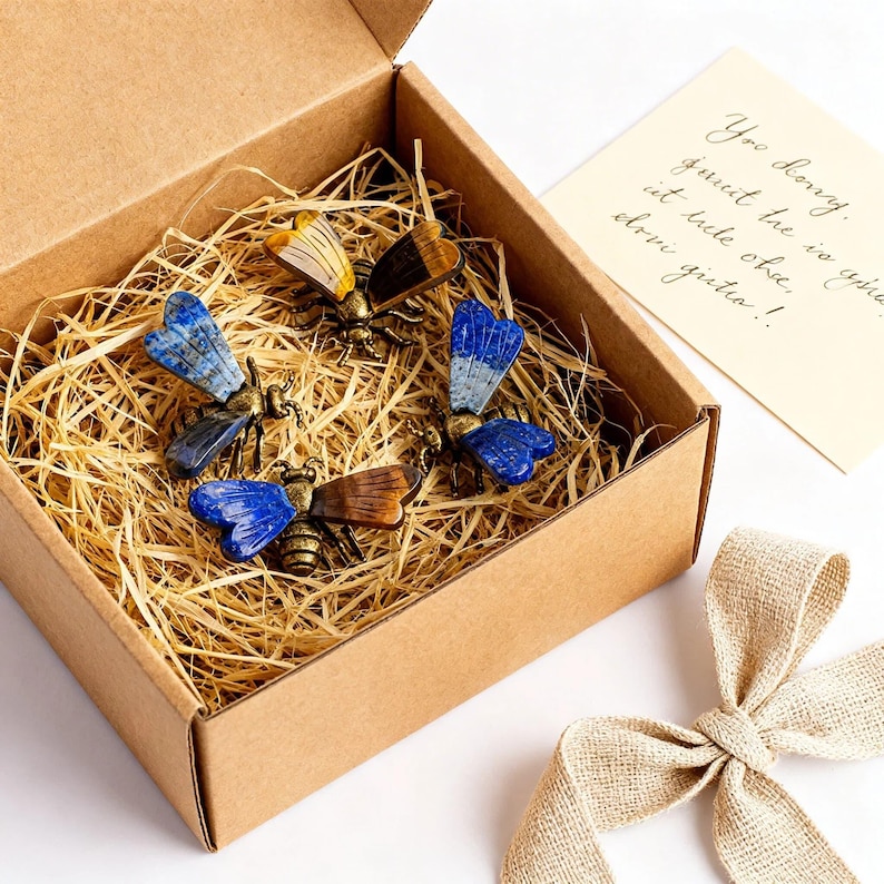 May include: Three bee-shaped ornaments with metallic bodies and wings crafted from blue lapis lazuli and brown tiger's eye gemstones, nestled in a brown cardboard gift box filled with straw, accompanied by a small card with handwritten text.