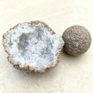 Op de afbeelding: Een gehalveerde geode met een sprankelend interieur van lichtblauwe kristallen. Daarnaast een ronde, grijze steen. De buitenste schil van de geode is ruw, getextureerd en bruin. De kristallen binnenin zijn samengeklonterd.