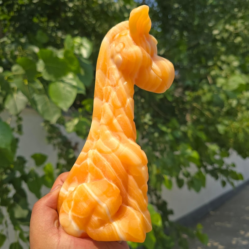 May include: A carved orange and white stone figurine in the shape of a giraffe. The sculpture features a smooth, polished surface with natural banded patterns in shades of translucent orange and opaque white, depicting the animal's neck and head.