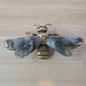 May include: A bee-shaped brooch featuring a metallic brass-toned body with segmented details and two large, translucent wings crafted from moss agate stone, displaying natural green and grey inclusions throughout the polished surfaces.