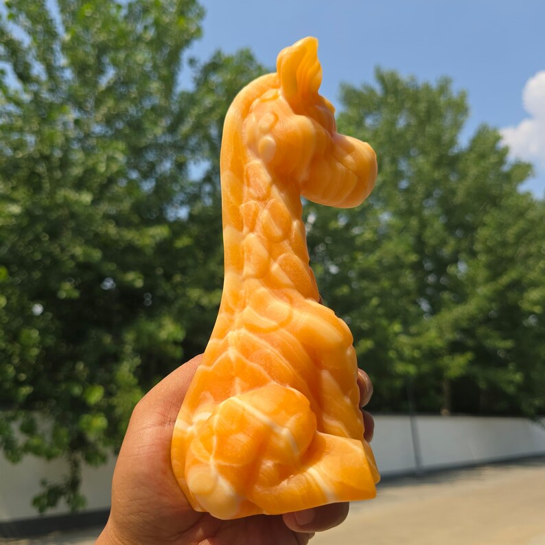 May include: A carved orange and white stone figurine shaped like a giraffe. The sculpture features a smooth, polished surface with natural swirling patterns in shades of bright orange and cream, depicting the animal in a seated position.