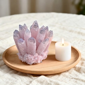 Peut inclure: Un amas de cristaux roses et violets sur un plateau en bois, à côté d'une bougie blanche allumée. Les cristaux ont un aspect givré. La bougie est cylindrique.