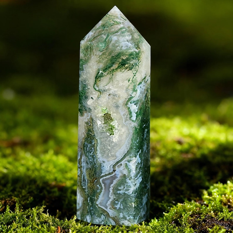 May include: A tall, polished moss agate crystal point. The crystal features a mix of translucent white and green hues, with intricate patterns resembling moss. The top is a pointed shape. The crystal is displayed on a bed of green moss.