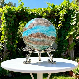 May include: A polished spherical stone featuring swirling patterns of vibrant turquoise, deep brown, and white mineral veins. The sphere rests securely on a silver-toned metal display stand with three ornate, curved legs and intricate scrollwork details.