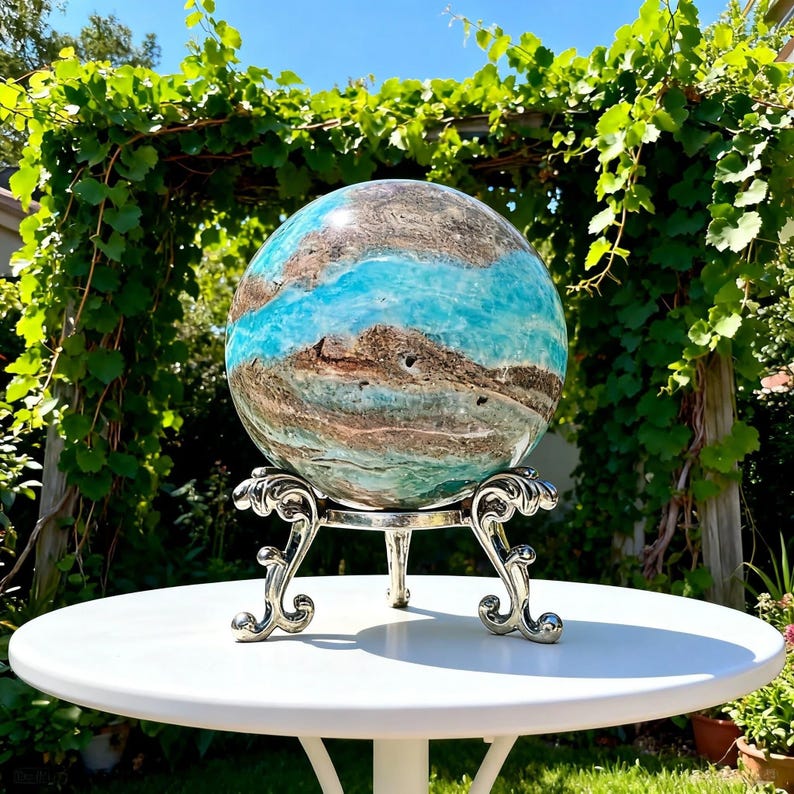 May include: A polished spherical stone featuring swirling patterns of vibrant turquoise, deep brown, and white mineral veins. The sphere rests securely on a silver-toned metal display stand with three ornate, curved legs and intricate scrollwork details.