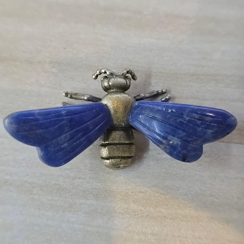 May include: A bee-shaped brooch featuring a metallic, antique-gold colored body with segmented details and two large, smooth, deep blue stone wings extending horizontally from the center. The insect design includes small antennae and legs.