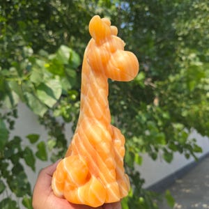 May include: A carved orange stone figurine in the shape of a giraffe. The sculpture features a smooth, polished surface with natural white banding and translucent layers, depicting the long neck, head, and body of the animal in a seated pose.