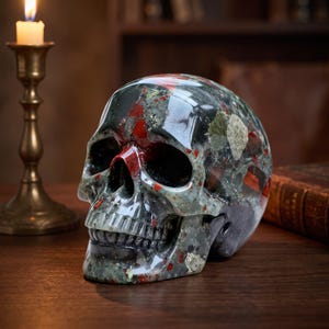 May include: A polished stone skull carving featuring a mottled pattern of dark green, deep red, and light gray mineral inclusions. The surface is smooth and reflective, showcasing the natural variations and textures of the stone material.