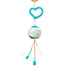 Op de afbeelding: Een goudkleurige sleutelhanger met een turquoise hartvormige bedel, heldere kristallen kralen en een ronde hanger met een berglandschap met kersenbloesems. De sleutelhanger heeft turquoise en heldere druppelvormige bedels.