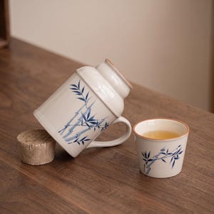 Puede incluir: Un juego de té de cerámica blanca con diseños de bambú azul. El juego incluye una taza grande, una taza pequeña con té y un posavasos de corcho. La taza está inclinada y la taza está en posición vertical, ambas sobre una superficie de madera.