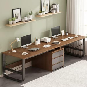 May include: A large, brown wooden desk with a metal frame, featuring two computer monitors, a laptop, and a three-drawer cabinet. The desk is set against a light green wall with floating shelves holding plants and framed artwork. A white rug is on the floor.