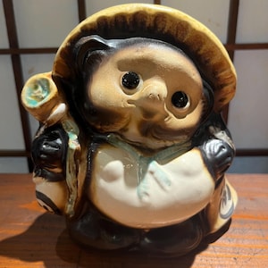 Peut inclure: Une figurine en céramique représentant un tanuki avec un chapeau de paille à larges bords, de grands yeux noirs et un ventre blanc. La figurine tient une bouteille de saké aux reflets verts et une pochette ronde, avec un émail brun, crème et bleu sarcelle.