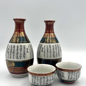 以下が含まれることがあります： 2つの装飾ボトルと2つのマッチングカップを備えた日本の酒セット。ボトルはマルーンのトップ、金のアクセント、黒と緑のバンドが付いています。カップとボトルは日本の書道で装飾されています。