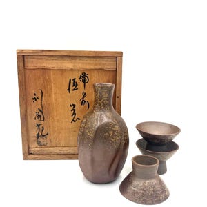 May include: A brown ceramic sake set with a bottle and four cups. The bottle has a rounded body and a narrow neck. The cups are small and round. A wooden box with Japanese characters is in the background.