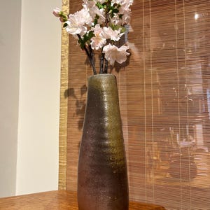 Puede incluir: Un jarrón de cerámica artesanal con degradado de tonos marrón y verde, que contiene un ramo de flores de cerezo blancas y rosas. El jarrón se exhibe sobre una superficie de madera. Mide unos 30 cm de alto.