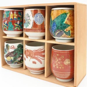 Puede incluir: Un juego de seis tazas de té japonesas de cerámica con varios diseños pintados a mano, incluyendo figuras, paisajes y patrones abstractos. Las tazas se exhiben en una caja de madera con compartimentos individuales. Los colores son rojo, verde, azul y dorado.